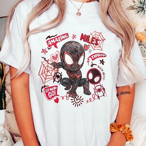 Op de afbeelding: Wit T-shirt met een cartoon illustratie van een superheld in een zwart en rood pak. Het ontwerp bevat de woorden "AMAZING", "MILES", "THWIP", "WEB-HEAD" en "GO-WEBS-GO!" samen met spinnenwebben en harten.