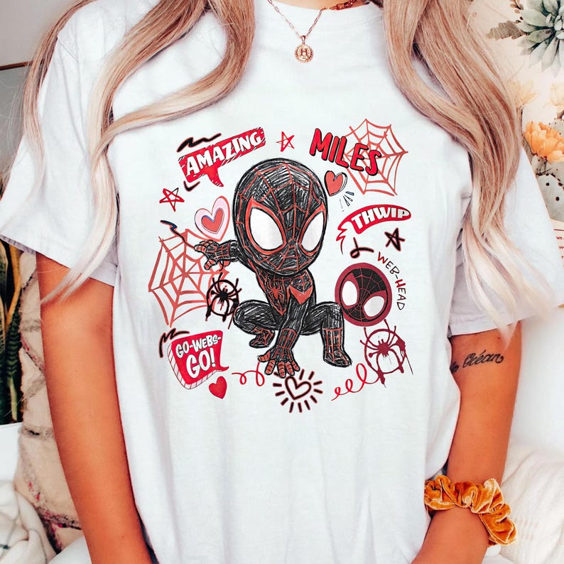 Op de afbeelding: Wit T-shirt met een cartoon illustratie van een superheld in een zwart en rood pak. Het ontwerp bevat de woorden "AMAZING", "MILES", "THWIP", "WEB-HEAD" en "GO-WEBS-GO!" samen met spinnenwebben en harten.