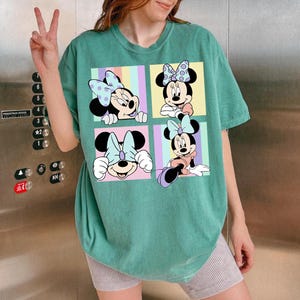 May include: A teal-colored t-shirt featuring a four-panel graphic of Minnie Mouse. Each panel showcases a different pose of Minnie, with pastel-colored backgrounds and polka-dot bows. The shirt has a relaxed fit.