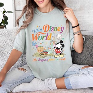 以下が含まれることがあります： ウォルト・ディズニー・ワールドのカラフルなグラフィックが特徴の、淡い緑色のTシャツ。デザインにはミッキーマウス、お城、そして「The happiest place on earth」というフレーズが含まれています。