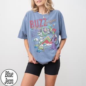 May include: A light blue t-shirt featuring a graphic of Buzz Lightyear from Toy Story. The design includes the words "BUZZ lightyear" and "LASER BLAST" in a colorful, cartoon style. The shirt is paired with black shorts.