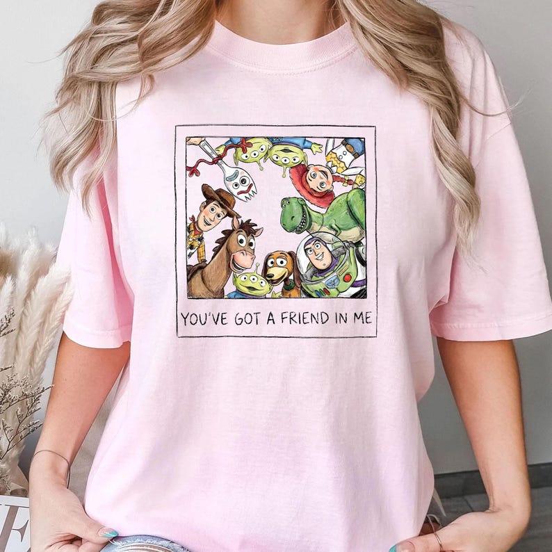 May include: Light pink t-shirt featuring a framed graphic of Toy Story characters, including Woody, Buzz Lightyear, and others. The text "YOU'VE GOT A FRIEND IN ME" is printed below the image. The shirt is made of a soft material.