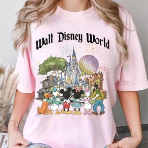 May include: A light pink t-shirt featuring a colorful graphic of Disney characters and landmarks. The text "Walt Disney World" is arched across the top. The graphic includes Mickey and Minnie Mouse, Donald Duck, Goofy, and a train.
