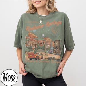 Pu&ograve; includere: T-shirt verde oliva con grafica di Radiator Springs, con testo "California, Radiator Springs, Welcome to Radiator Springs." Il design include auto, un serbatoio d'acqua e paesaggio desertico.