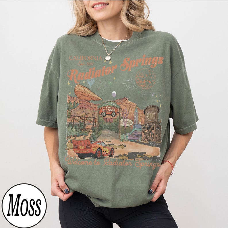 Pu&ograve; includere: T-shirt verde oliva con grafica di Radiator Springs, con testo "California, Radiator Springs, Welcome to Radiator Springs." Il design include auto, un serbatoio d'acqua e paesaggio desertico.