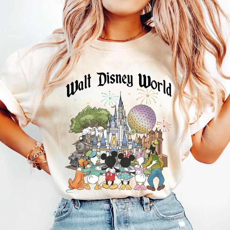 May include: Cream-colored t-shirt featuring a colorful graphic of Disney characters in front of Cinderella's castle, Spaceship Earth, and a train. The words "Walt Disney World" are printed above the graphic in a black, stylized font. The shirt has short sleeves.