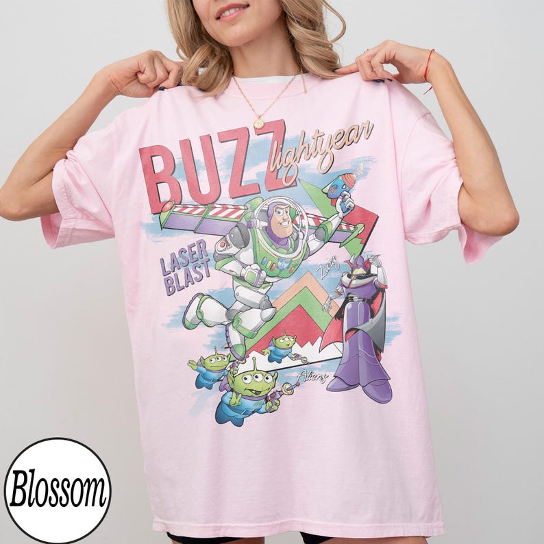 May include: A light pink t-shirt featuring a graphic of Buzz Lightyear and other characters from the movie Toy Story. The text "BUZZ lightyear" and "LASER BLAST" are printed on the front. The shirt has short sleeves and a relaxed fit.