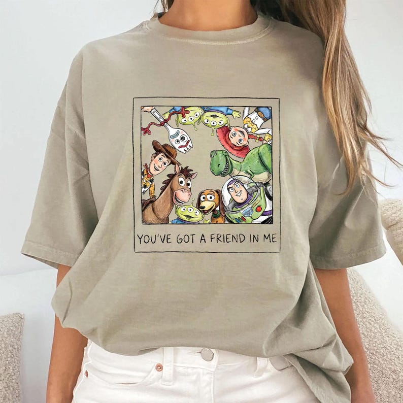May include: A light olive green t-shirt featuring a framed cartoon illustration of characters from the movie Toy Story. The illustration includes Woody, Buzz Lightyear, and other characters. The text "YOU'VE GOT A FRIEND IN ME" is below the illustration.