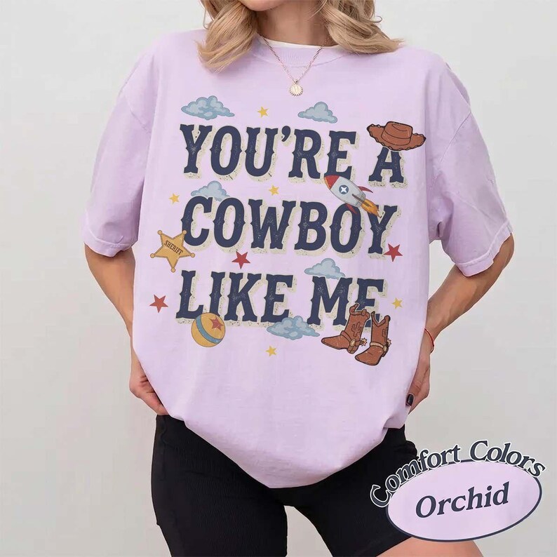 May include: A light purple short-sleeve t-shirt featuring the dark blue text "YOU'RE A COWBOY LIKE ME" surrounded by graphic illustrations of a rocket, a sheriff star, a cowboy hat, a ball, cowboy boots, clouds, and small stars.