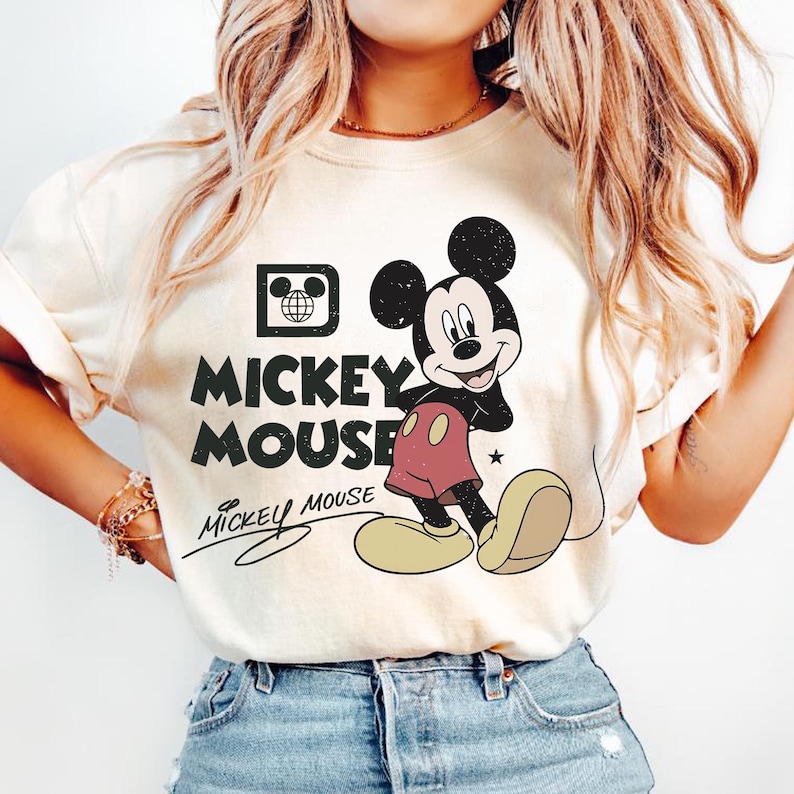 Op de afbeelding: Cr&egrave;mekleurig T-shirt met een afbeelding van Mickey Mouse. Het ontwerp bevat Mickey Mouse in zwart, rood en geel, met de woorden "Mickey Mouse" in een verweerde lettertype. Een kleine ster is ook in het ontwerp opgenomen.