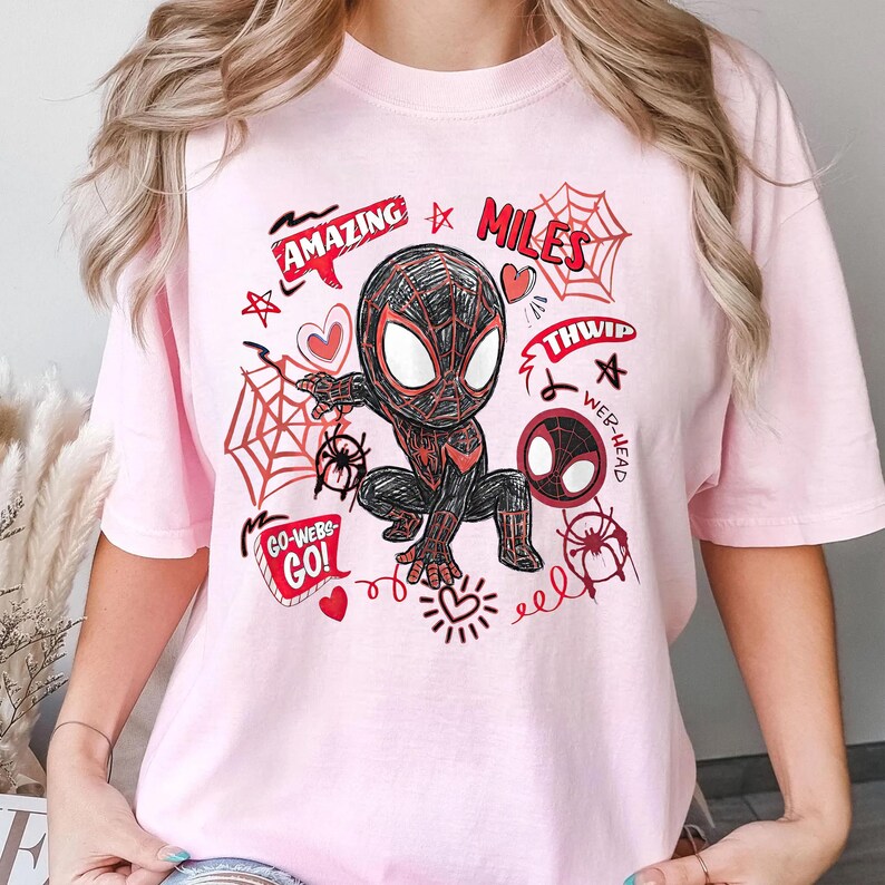 Op de afbeelding: Lichtroze T-shirt met een cartoon illustratie van Spider-Man in een gehurkte positie. Het ontwerp bevat rode en zwarte accenten, spinnenwebben en de woorden "Amazing", "Miles", "Go-Webs-Go!" en "Thwip".