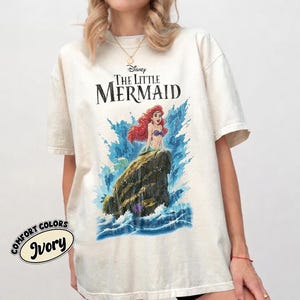 May include: An ivory short-sleeve t-shirt featuring a graphic print of Ariel from The Little Mermaid sitting on a rock amidst blue ocean waves. The text "Disney The Little Mermaid" is printed in black above the character. A small circular logo reads "Comfort Colors Ivory" in the bottom left corner.