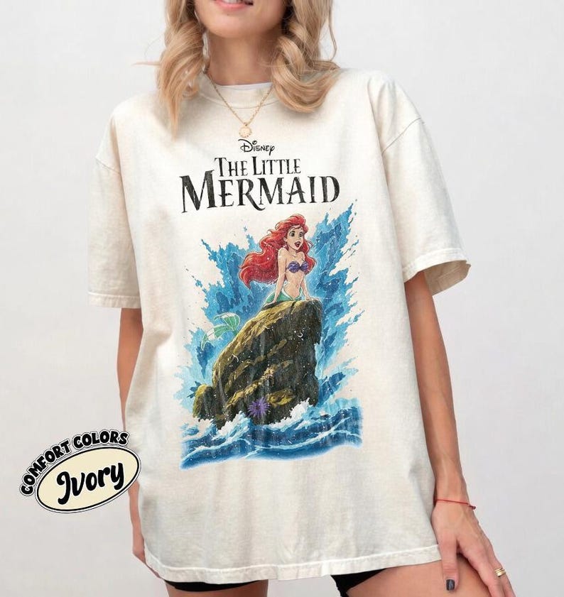 May include: An ivory short-sleeve t-shirt featuring a graphic print of Ariel from The Little Mermaid sitting on a rock amidst blue ocean waves. The text "Disney The Little Mermaid" is printed in black above the character. A small circular logo reads "Comfort Colors Ivory" in the bottom left corner.