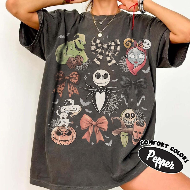 May include: A charcoal grey t-shirt featuring a graphic print of characters and motifs from The Nightmare Before Christmas, including Jack Skellington, Sally, Zero, Oogie Boogie, bows, spiderwebs, and bats. The text Comfort Colors Pepper is visible.