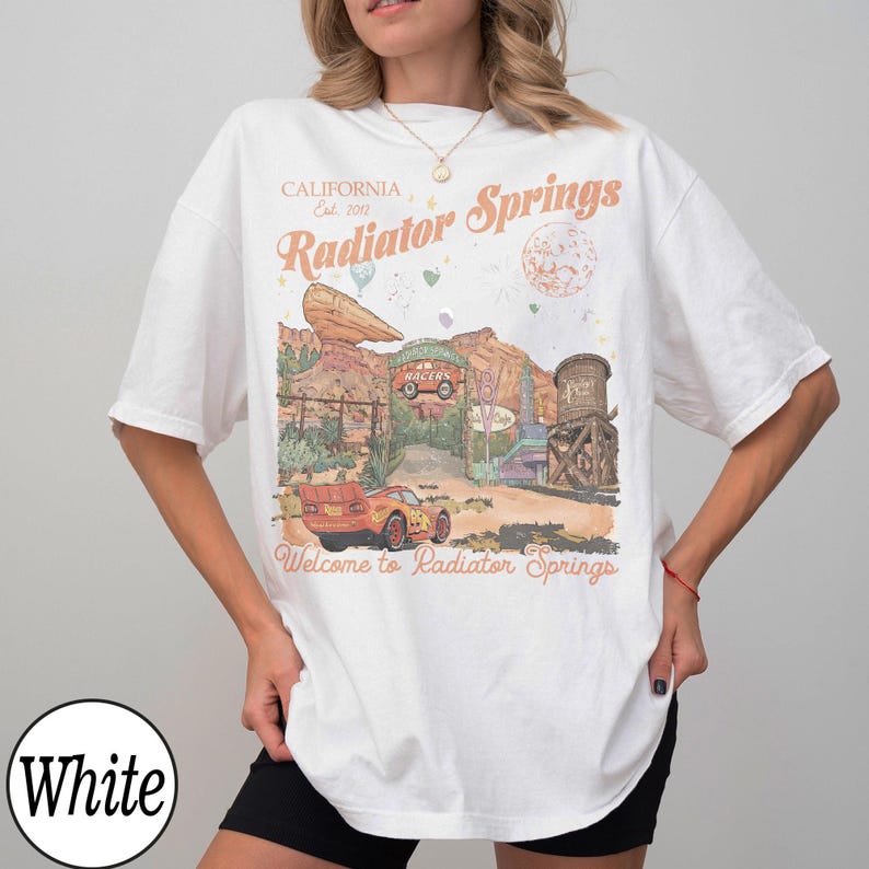 Pu&ograve; includere: T-shirt bianca con grafica di Radiator Springs, con testo che recita "California Est. 2012" e "Welcome to Radiator Springs." Il design include un'auto dei cartoni animati, un paesaggio desertico e un serbatoio d'acqua.
