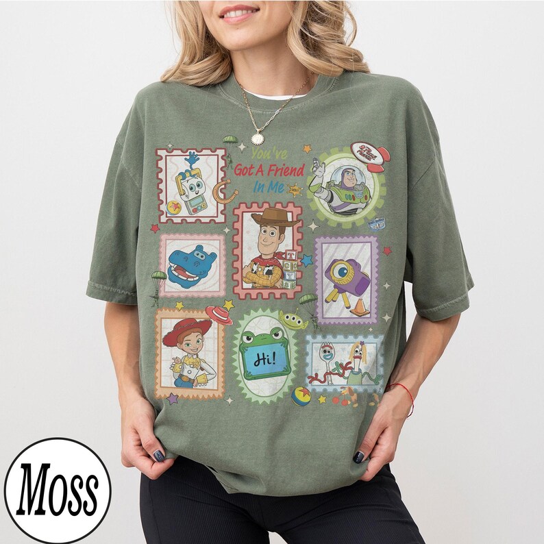 May include: A moss green short-sleeve t-shirt featuring a graphic print of various Toy Story characters in postage stamp-style frames. The text "You've Got A Friend In Me" is printed in the center, surrounded by illustrations of Woody, Buzz Lightyear, Jessie, and other characters.