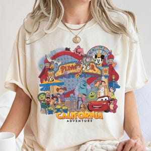 May include: Cream-coloured t-shirt with a vibrant graphic of the Pixar California Adventure theme park. The design showcases various Pixar characters, the Pixar logo, and the words "CALIFORNIA ADVENTURE".