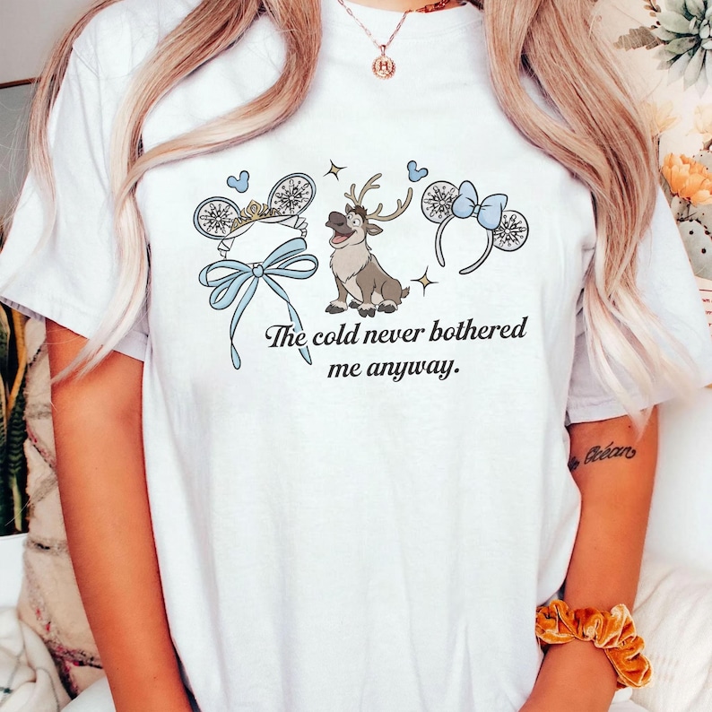 May include: White t-shirt with a cartoon design including a reindeer, mouse ears, and a bow in blue and white. The text "The cold never bothered me anyway" is also featured on the shirt.