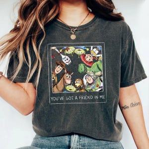 May include: Dark gray t-shirt featuring a colorful graphic of Toy Story characters within a white rectangular frame. The text "YOU'VE GOT A FRIEND IN ME" is printed below the graphic. The shirt has a crew neck and short sleeves.