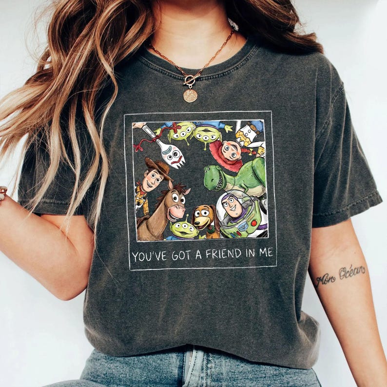 May include: Dark gray t-shirt featuring a colorful graphic of Toy Story characters within a white rectangular frame. The text "YOU'VE GOT A FRIEND IN ME" is printed below the graphic. The shirt has a crew neck and short sleeves.