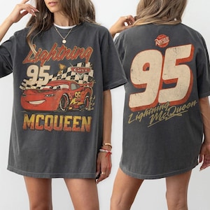 May include: Dark gray t-shirt featuring a graphic of Lightning McQueen from the Cars movie. The front design includes the number 95, a checkered flag, and the name McQueen. The back of the shirt displays a large number 95 and the Lightning McQueen name.
