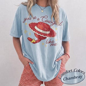 May include: A light blue short-sleeve t-shirt featuring a graphic of a red cowgirl hat with a braided ponytail and the text "you're a Cowgirl like me." The design includes small yellow star accents. The shirt is made by Comfort Colors.