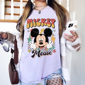 May include: A light purple t-shirt featuring a graphic of Mickey Mouse with the text "Mickey Mouse". The design includes a retro font, stars, and lightning bolts. Mickey is making a peace sign with both hands.