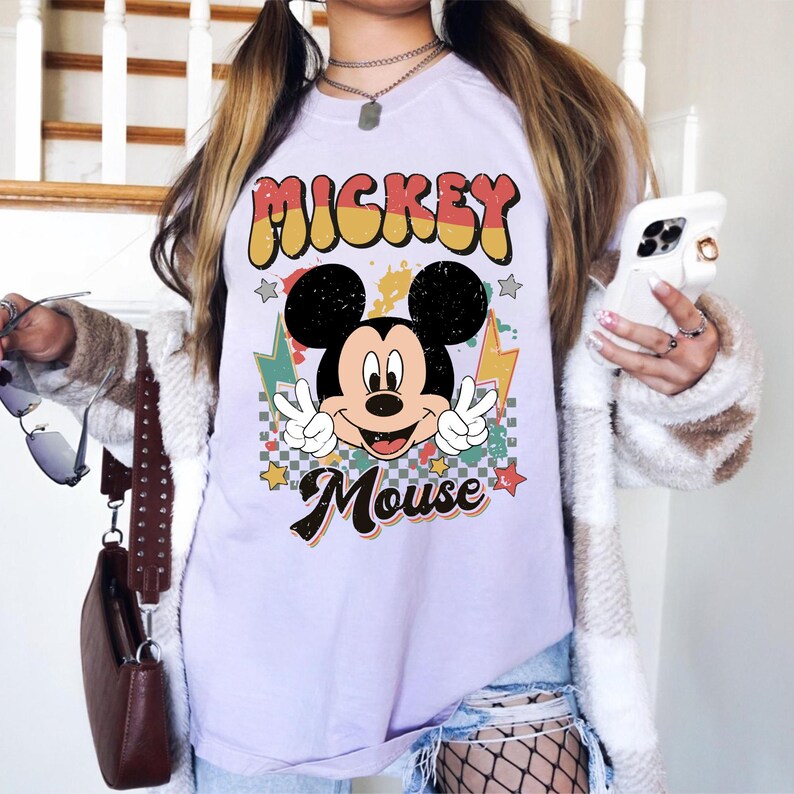 May include: A light purple t-shirt featuring a graphic of Mickey Mouse with the text "Mickey Mouse". The design includes a retro font, stars, and lightning bolts. Mickey is making a peace sign with both hands.