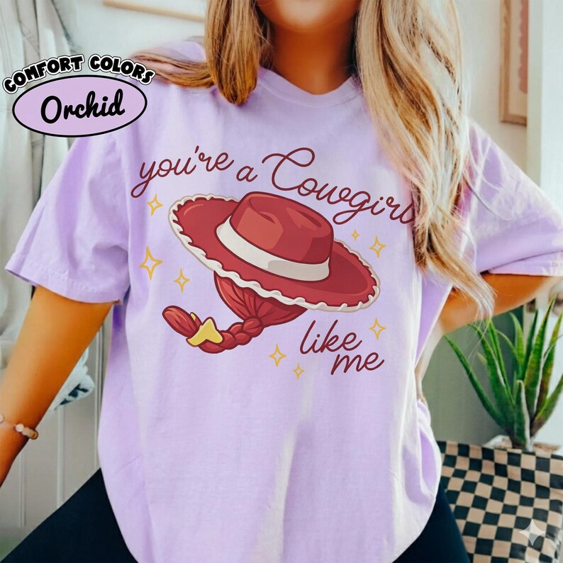 May include: A light purple t-shirt featuring a graphic of a red cowgirl hat with a braided ponytail and the text "you're a Cowgirl like me" in dark red script. The upper left corner includes a circular label reading "COMFORT COLORS Orchid".