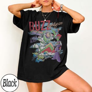 May include: Black t-shirt featuring a graphic of Buzz Lightyear from Toy Story. The design includes the text "BUZZ lightyear" and "LASER BLAST", along with images of Buzz, aliens, and Zurg. The shirt is a relaxed fit.