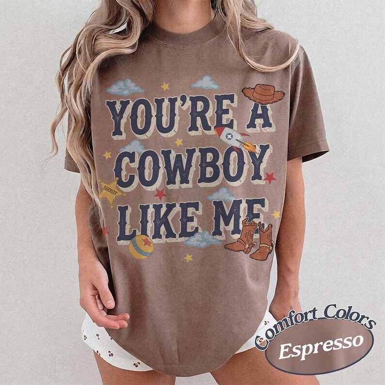 May include: A brown short-sleeve t-shirt featuring the text "YOU'RE A COWBOY LIKE ME" in dark blue letters. The design includes illustrations of a cowboy hat, a rocket, a sheriff star, a ball, and cowboy boots, surrounded by small clouds and stars.