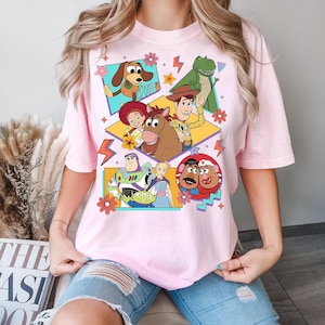 Puede incluir: Camiseta rosa claro con un dise&ntilde;o gr&aacute;fico colorido de personajes de la pel&iacute;cula animada Toy Story. El dise&ntilde;o incluye a Woody, Buzz Lightyear, Jessie y otros personajes en un estilo retro. La camiseta tiene cuello redondo y mangas cortas.