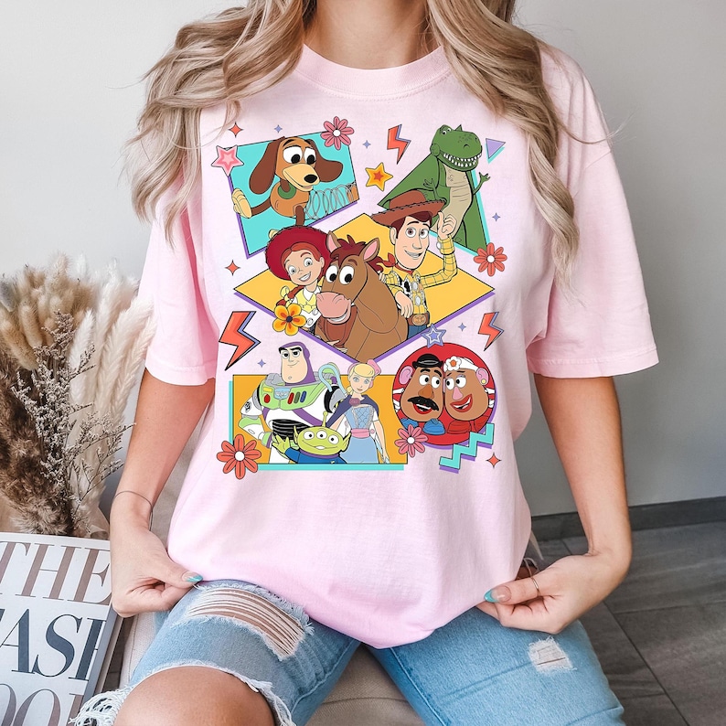 Puede incluir: Camiseta rosa claro con un dise&ntilde;o gr&aacute;fico colorido de personajes de la pel&iacute;cula animada Toy Story. El dise&ntilde;o incluye a Woody, Buzz Lightyear, Jessie y otros personajes en un estilo retro. La camiseta tiene cuello redondo y mangas cortas.