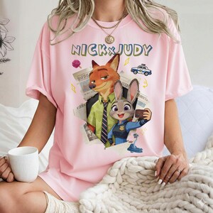 May include: Pink t-shirt featuring the text "NICK & JUDY" above a graphic of Nick Wilde and Judy Hopps from Zootopia. Nick is wearing a green tie and Judy is taking a selfie. A police car and newspaper clippings are also featured.
