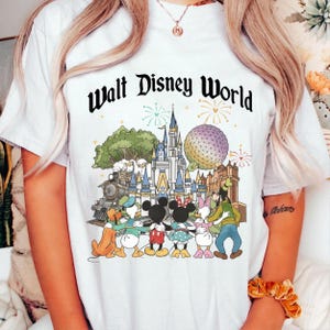 May include: White t-shirt featuring a colorful graphic of Disney characters and landmarks, including a castle, train, and Spaceship Earth. The text "Walt Disney World" is printed in black script above the image.