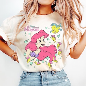 May include: Cream-colored t-shirt featuring a graphic of Ariel from Disney's The Little Mermaid. Ariel has pink hair and a purple seashell top. The design includes Flounder, seahorses, and bubbles. The text "The Little Mermaid" is above Ariel.