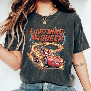 Może przedstawiać: Ciemnoszara koszulka z postacią Zygzaka McQueena z filmu Auta. Grafika przedstawia czerwony samoch&oacute;d wyścigowy z płomieniami i napisem "Lightning McQueen". Koszulka ma okrągły dekolt i kr&oacute;tkie rękawy.
