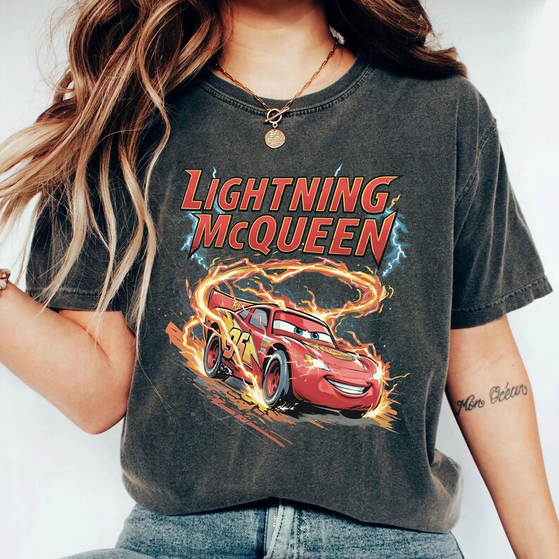 Może przedstawiać: Ciemnoszara koszulka z postacią Zygzaka McQueena z filmu Auta. Grafika przedstawia czerwony samoch&oacute;d wyścigowy z płomieniami i napisem "Lightning McQueen". Koszulka ma okrągły dekolt i kr&oacute;tkie rękawy.