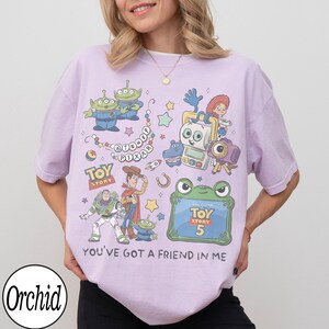 Puede incluir: Una camiseta de color morado claro con un estampado gr&aacute;fico colorido de personajes de Toy Story, incluyendo a Woody, Buzz Lightyear y alien&iacute;genas. El dise&ntilde;o incluye el texto Disney Pixar, Toy Story, Toy Story 5 y la frase You've Got A Friend In Me.