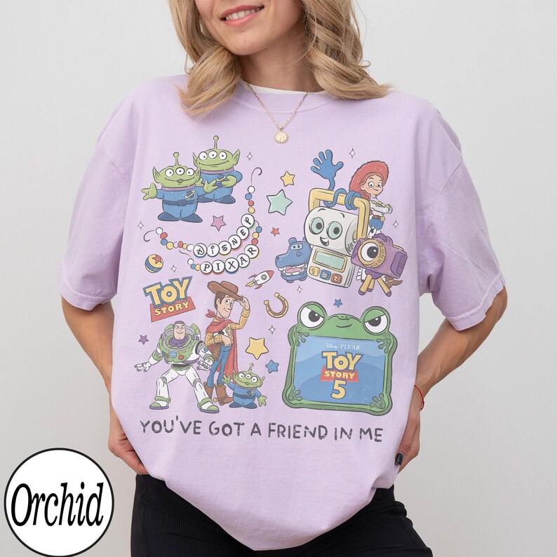 Puede incluir: Una camiseta de color morado claro con un estampado gr&aacute;fico colorido de personajes de Toy Story, incluyendo a Woody, Buzz Lightyear y alien&iacute;genas. El dise&ntilde;o incluye el texto Disney Pixar, Toy Story, Toy Story 5 y la frase You've Got A Friend In Me.