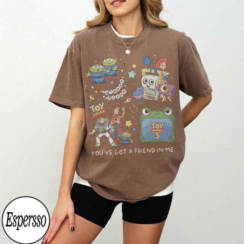 Puede incluir: Una camiseta marr&oacute;n de manga corta con un estampado gr&aacute;fico de personajes de Toy Story, incluyendo a Buzz Lightyear, Woody y alien&iacute;genas. El dise&ntilde;o incluye el texto Toy Story, Toy Story 5 y You've Got A Friend In Me, junto a estrellas.