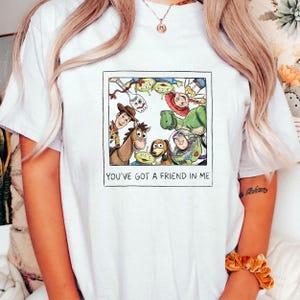 May include: White t-shirt featuring a framed illustration of characters from the animated film "Toy Story." The image includes Woody, Buzz Lightyear, and other characters. The text "YOU'VE GOT A FRIEND IN ME" is printed below the illustration.