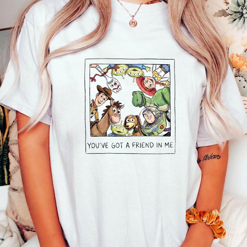 May include: White t-shirt featuring a framed illustration of characters from the animated film "Toy Story." The image includes Woody, Buzz Lightyear, and other characters. The text "YOU'VE GOT A FRIEND IN ME" is printed below the illustration.