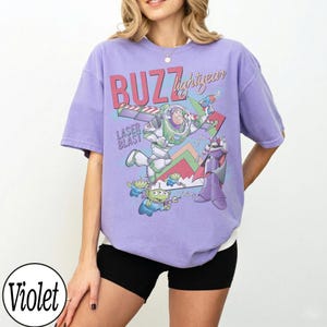 May include: Violet-colored t-shirt featuring a graphic of Buzz Lightyear with the text "BUZZ lightyear" and "LASER BLAST". The design includes cartoon characters and colorful accents. The shirt has short sleeves and a relaxed fit.