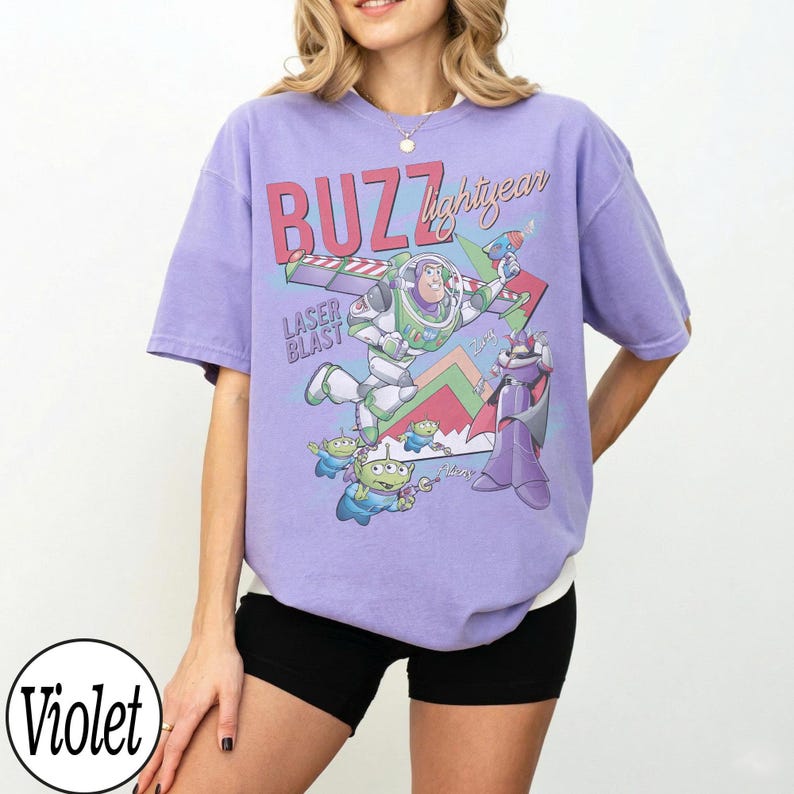 May include: Violet-colored t-shirt featuring a graphic of Buzz Lightyear with the text "BUZZ lightyear" and "LASER BLAST". The design includes cartoon characters and colorful accents. The shirt has short sleeves and a relaxed fit.