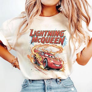 Może przedstawiać: Kremowa koszulka z napisem "LIGHTNING McQUEEN" w kolorze czerwonym i niebieskim oraz grafiką czerwonego samochodu wyścigowego. Projekt zawiera płomienie i błyskawice, tworząc dynamiczny i energiczny wygląd.