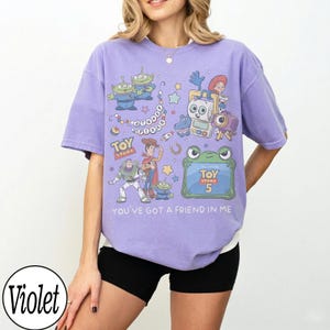 Puede incluir: Una camiseta violeta de manga corta con un estampado gr&aacute;fico de varios personajes de Toy Story, incluyendo a Woody, Buzz Lightyear y alien&iacute;genas. El dise&ntilde;o incluye el texto "Toy Story", "Toy Story 5" y "You've got a friend in me" en blanco.