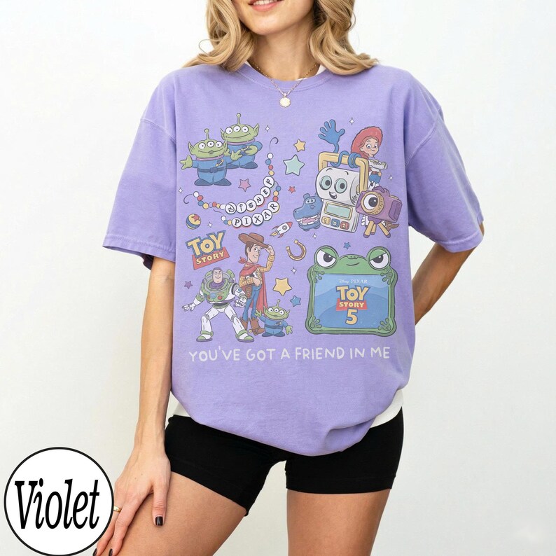 Puede incluir: Una camiseta violeta de manga corta con un estampado gr&aacute;fico de varios personajes de Toy Story, incluyendo a Woody, Buzz Lightyear y alien&iacute;genas. El dise&ntilde;o incluye el texto "Toy Story", "Toy Story 5" y "You've got a friend in me" en blanco.