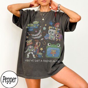 Puede incluir: Una camiseta de manga corta gris carb&oacute;n con un estampado gr&aacute;fico de varios personajes de Toy Story, incluidos Buzz Lightyear, Woody y los Aliens. El dise&ntilde;o incluye el texto Disney Pixar, Toy Story, Toy Story 5 y You've Got A Friend In Me.