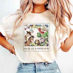 May include: Cream-colored t-shirt featuring a framed illustration of characters from the animated film "Toy Story." The image includes Woody, Buzz Lightyear, and other characters. The text "YOU'VE GOT A FRIEND IN ME" is printed below the image.
